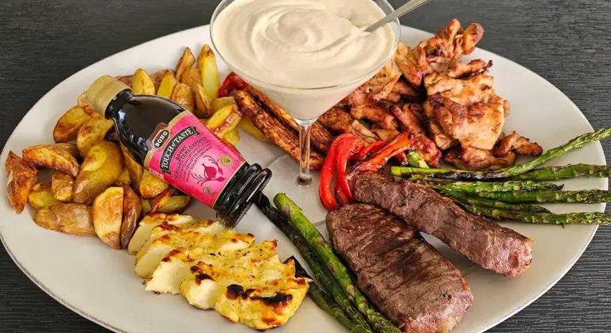 En festlig tallrik med grillad biff, kyckling, halloumi, potatis, sparris och en krämig sås. En flaska "Touch of Taste" sås till grillat står lutad mot potatisen.