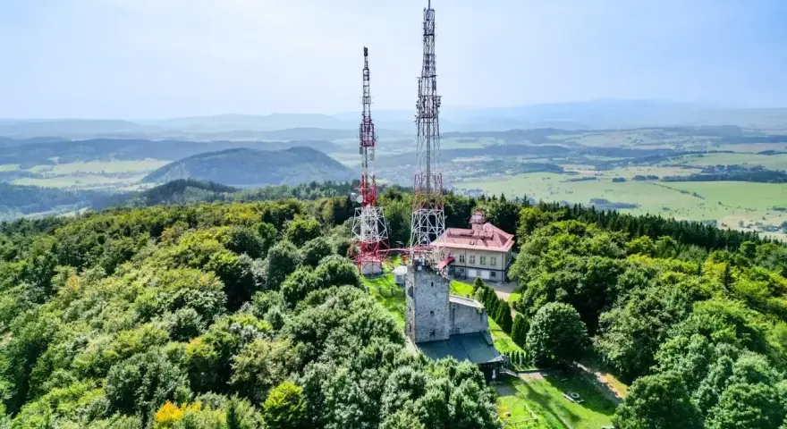 Wieże telekomunikacyjne na zalesionym szczycie Chełmiec. W tle rozciąga się malowniczy krajobraz pogórza.