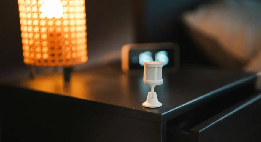 Sensor de movimiento blanco sobre mesita de noche, junto a lámpara y reloj. Ideal para saber como dejar fijo un sensor de movimiento.