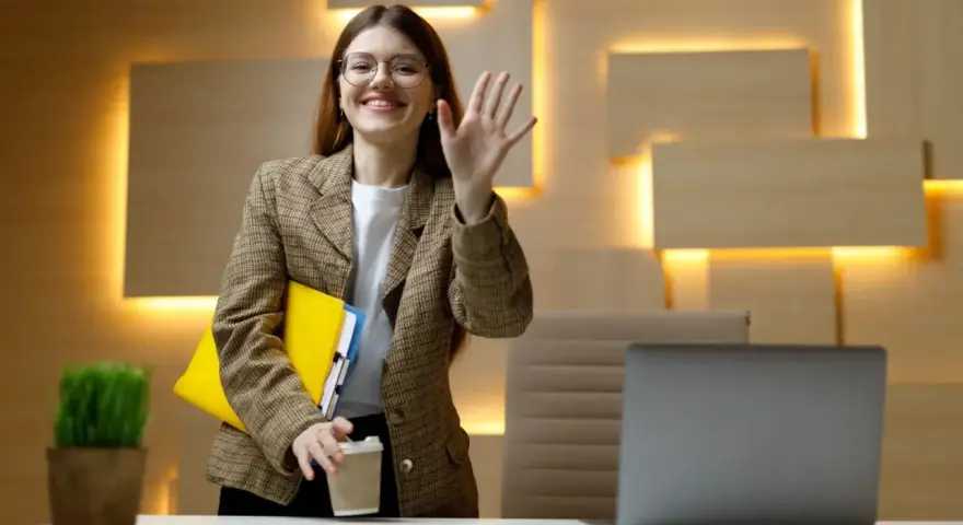 Uśmiechnięta kobieta w okularach macha ręką, trzymając teczkę i kawę. Czy dzień próbny jest płatny?