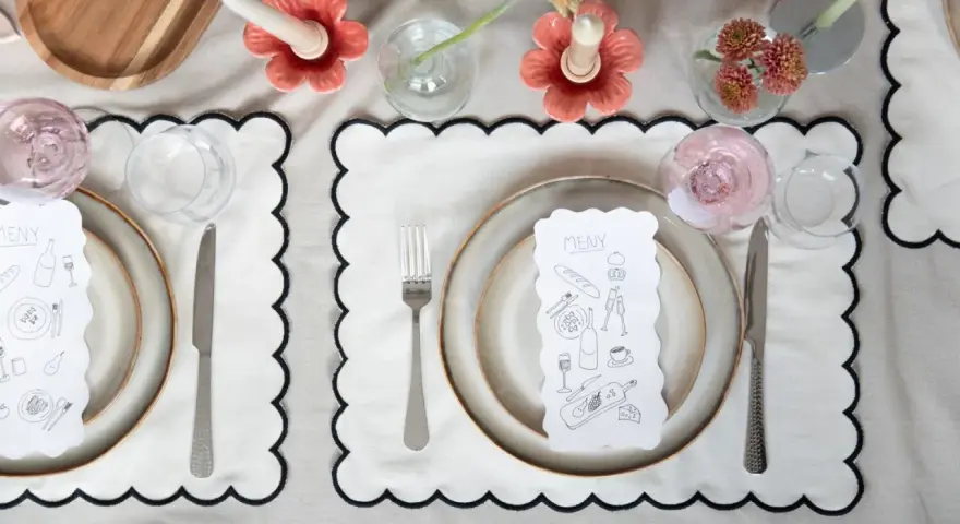 A table setting with a gold-colored tablecloth, white placemats with black scalloped edges, plates, cutlery, glasses, and a menu with illustrations.