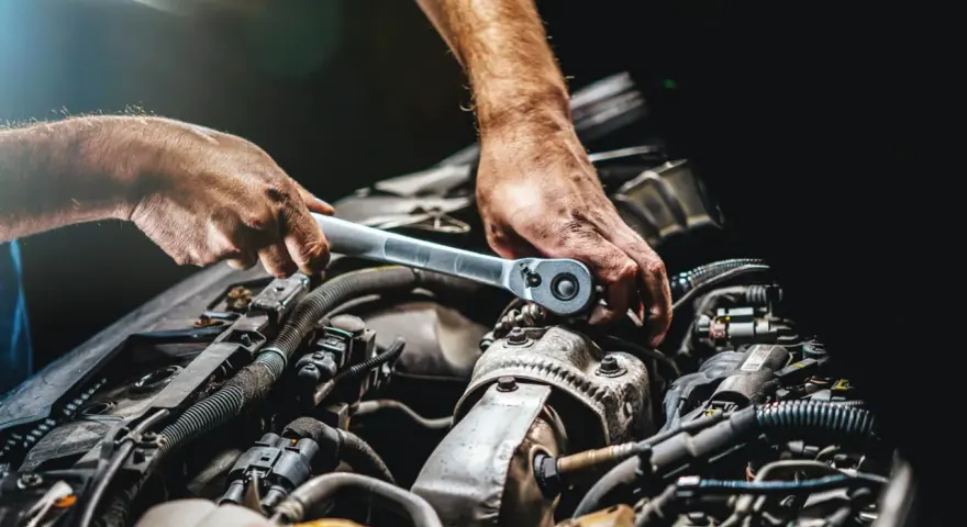 Mechanik dokręca śrubę kluczem w silniku samochodu. Widać jego dłonie i fragmenty silnika, być może podczas rutynowej kontroli lub naprawy.