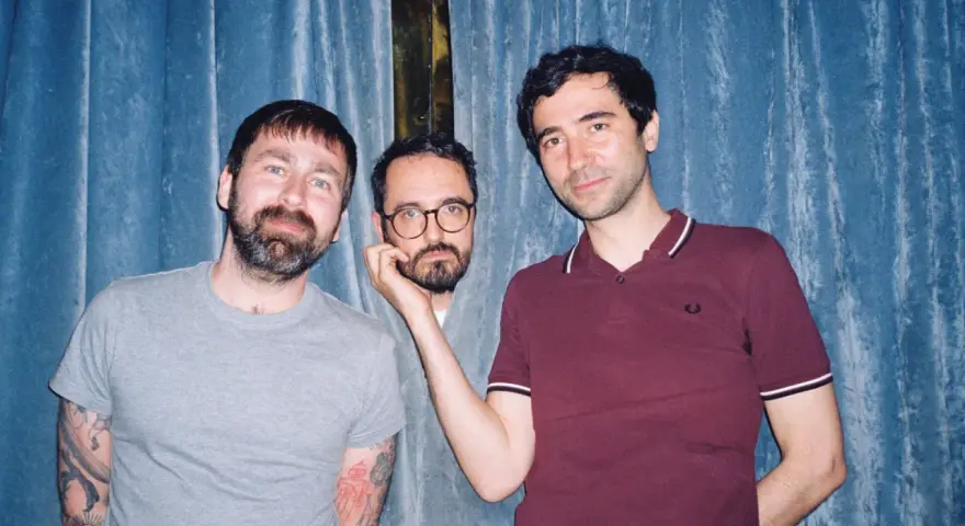 Trois hommes posent devant un rideau bleu. L'un d'eux, au centre, est un groupe musique espagnol année 2000.
