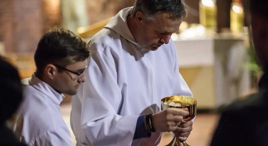 Szafarz, w białej albie, trzyma złoty kielich z hostiami. Obok niego młodszy mężczyzna w okularach.