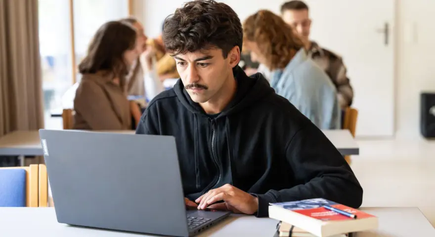 Ein junger Mann mit Schnurrbart konzentriert sich auf seinen Laptop, während er ein evangelisches Fernstudium Theologie absolviert.