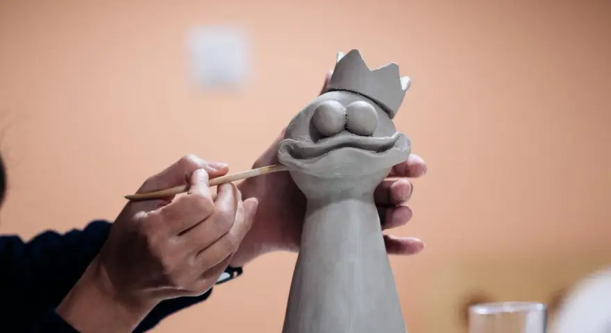 Manos trabajando con arcilla para crear una figura de rana con corona.