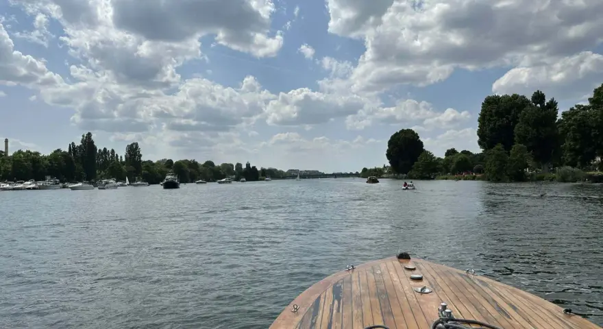 Blick vom Bug eines Holzbootes auf einen Fluss mit vielen Booten und grünen Ufern. Ein schöner Tag, fast wie ein Heidelberg See.