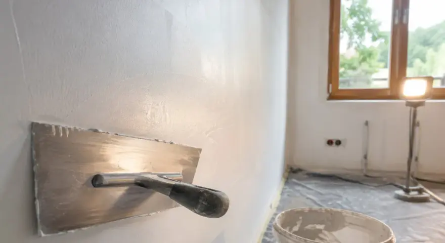 Nakładanie gładzi na ścianę za pomocą kielni. W tle okno i lampa budowlana.