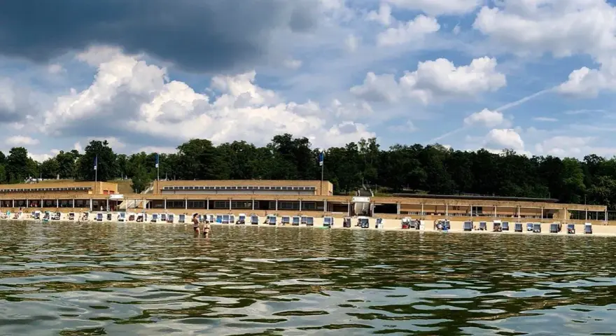 Das Strandbad Wannsee mit seiner langen Promenade und vielen Liegestühlen, im Hintergrund grüne Bäume und ein bewölkter Himmel.