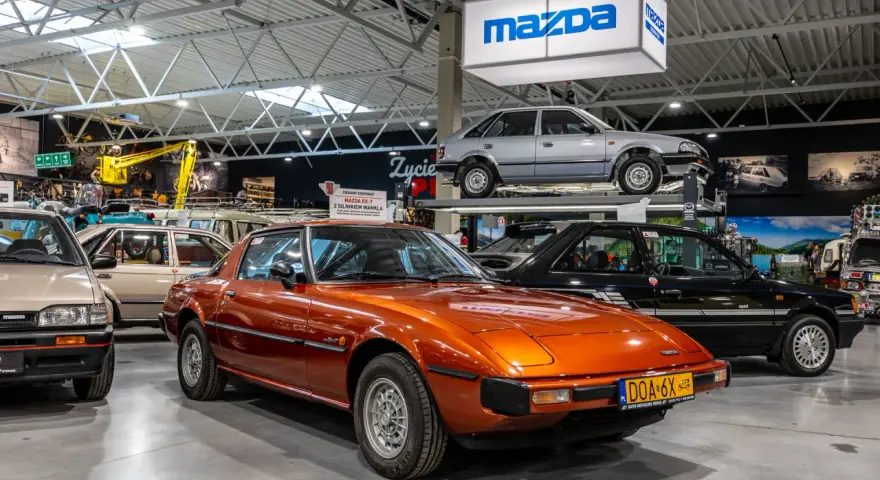 Pomarańczowa Mazda RX-7 błyszczy w muzeum samochodów, obok innych klasyków marki.