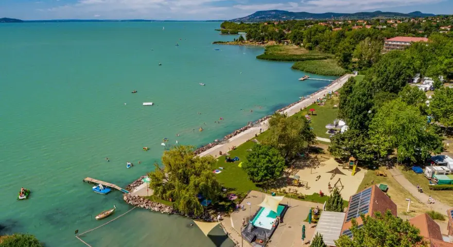 Słoneczny dzień na kempingu Balaton. Ludzie kąpią się w jeziorze i basenie, bawią na placu zabaw, a kampery stoją wśród drzew.