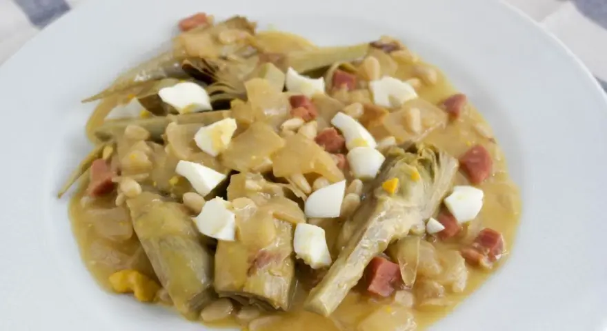 Plato de alcachofas con jamón, huevo duro y piñones, servido en una cazuela.