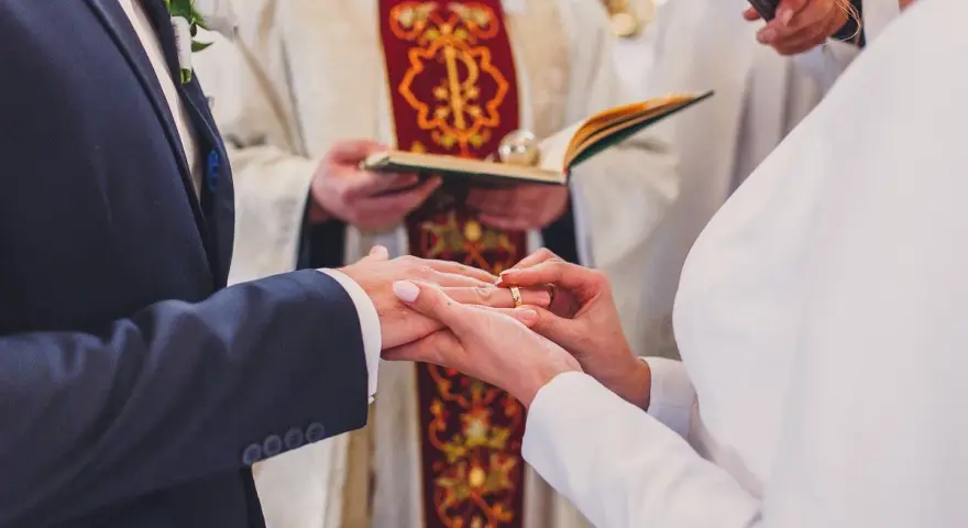 Ceremonia ślubna w kościele. Młoda para wymienia obrączki. W tle ksiądz z księgą. Warto wiedzieć, ile kosztuje ślub kościelny.