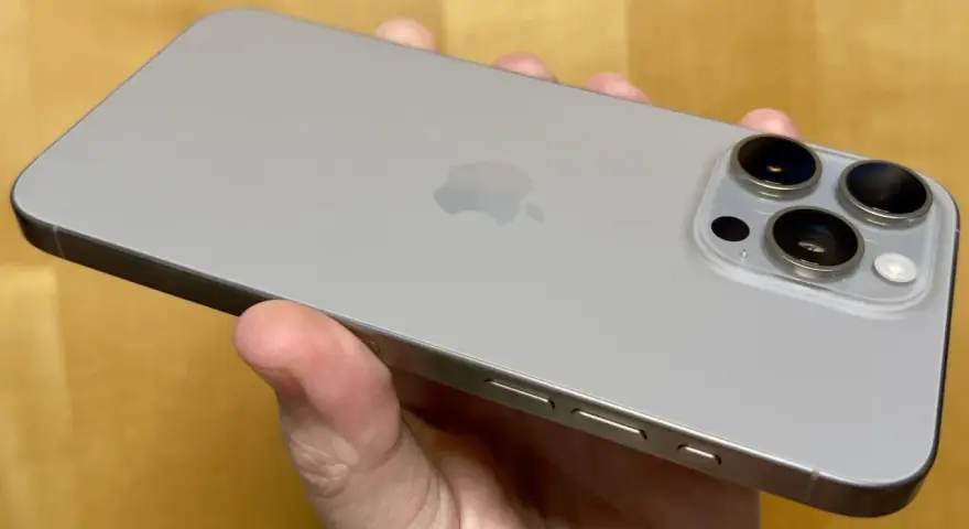 En hand håller en silverfärgad iPhone 15 Pro Max. Kameramodulen med tre linser syns tydligt.