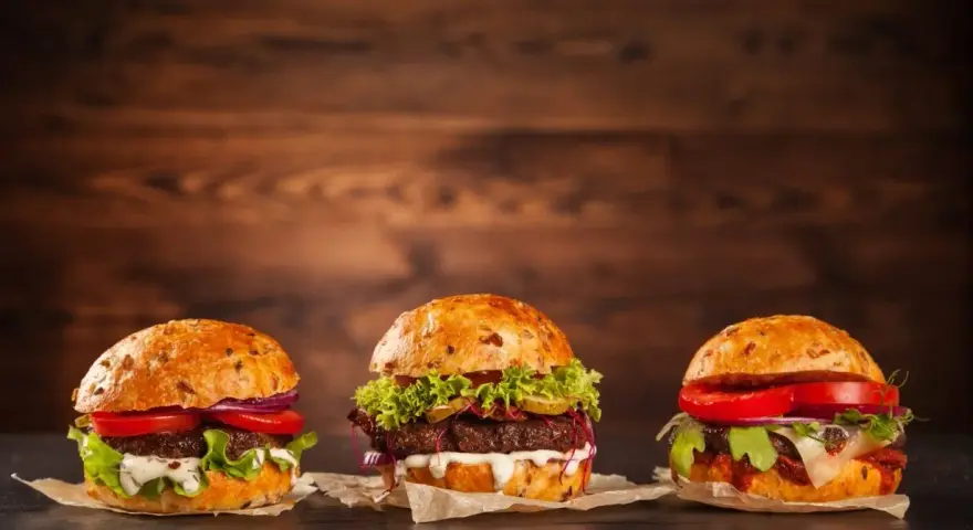 Trzy apetyczne burgery z różnymi dodatkami na drewnianym tle.