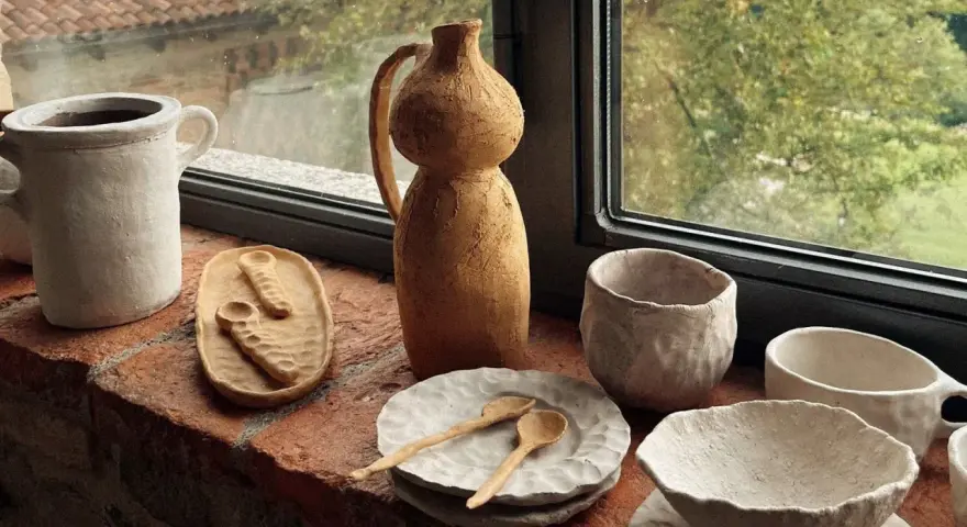 Ceramica fatta in casa: vasi, ciotole e cucchiaini grezzi, posati su un davanzale di mattoni, con vista su un giardino verde.