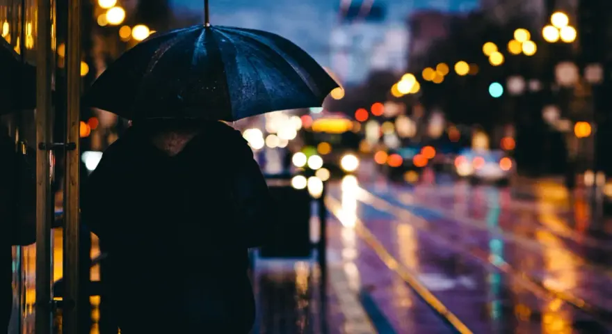 Person sous un parapluie noir, attendant sur un trottoir mouillé. Les lumières de la ville se reflètent sur la chaussée, créant de belles photos de pluie.