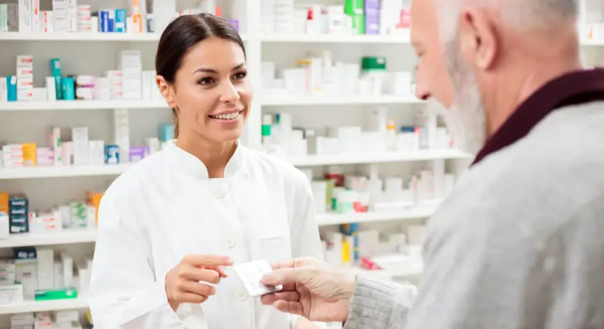 Uśmiechnięta farmaceutka podaje starszemu panu leki. W tle apteczne półki pełne leków, w tym poltixa.
