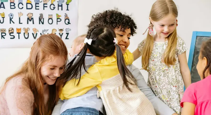 Uśmiechnięty czterolatek przytula się do nauczycielki, otoczony przez inne dzieci. W tle wisi plakat z literami i zwierzątkami.