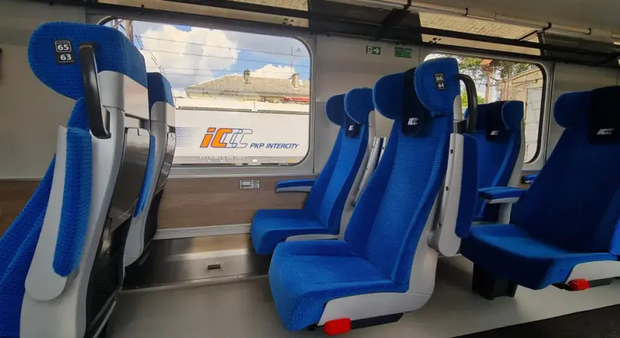 Wygodne, niebieskie fotele w wagonach PKP Intercity bez przedziałów. Widok na mijany krajobraz i inne wagony.