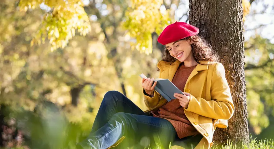 Mujer con boina roja lee un libro bajo un árbol, inspirada por los temas de cuentos del otoño.