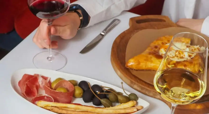 Ręka trzyma kieliszek czerwonego wina wytrawnego. Obok talerz z wędliną, oliwkami i grissini oraz chaczapuri i kieliszek białego wina.