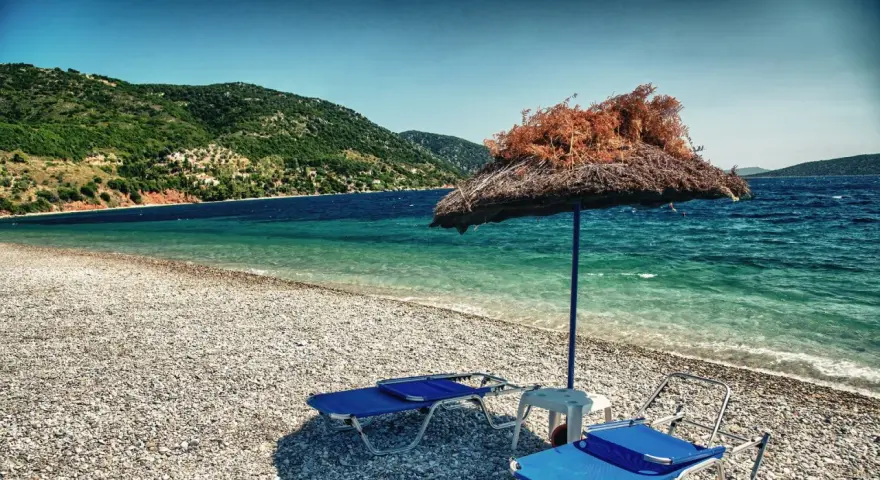 Plaża z leżakami i parasolem, w tle zielone wzgórza i turkusowe morze. To idealne miejsce na wypoczynek na grupie wysp na Morzu Egejskim.