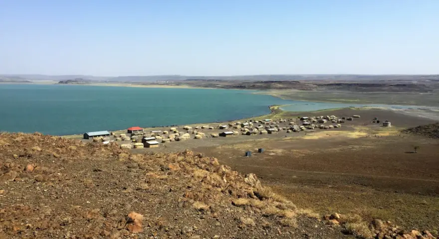 Widok na osadę nad turkusowym jeziorem Turkana. W oddali widać górzysty krajobraz.
