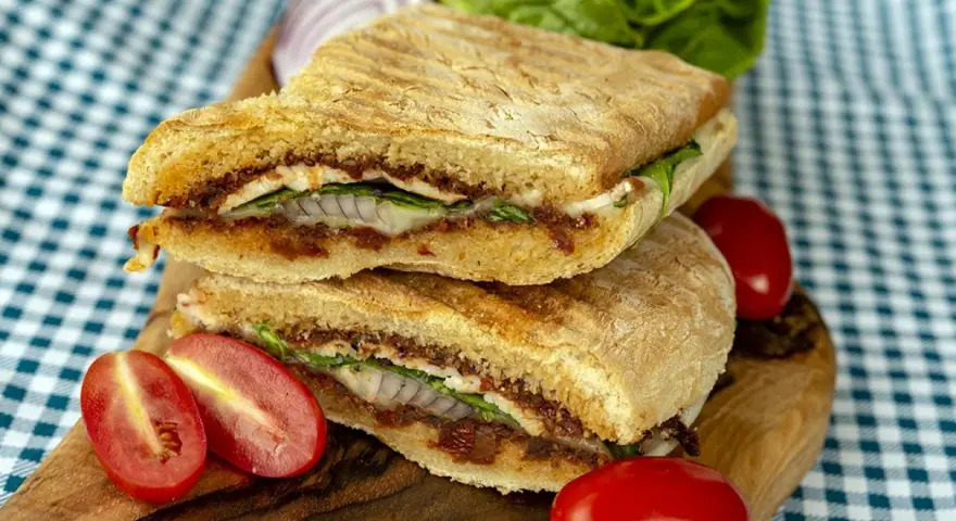Dwie połówki panini z serem, suszonymi pomidorami, rukolą i cebulą na desce, obok pomidorków i sałaty.