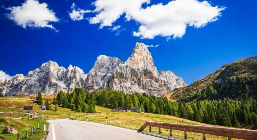 Strada panoramica per esplorare i passi del Trentino in moto, con imponenti vette dolomitiche sullo sfondo e un cielo azzurro con nuvole bianche.
