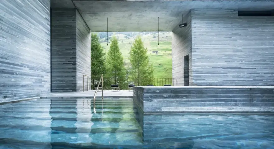 Piscine intérieure des thermes de Vals Zumthor, avec vue sur les montagnes verdoyantes et les sapins. L'eau bleue reflète les murs en pierre grise.