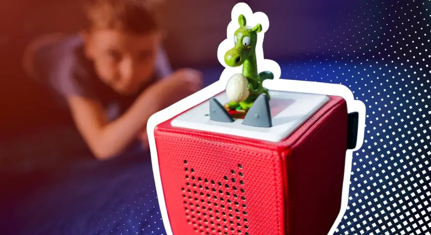 Ein grüner Drache mit Ei auf einer roten Toniebox, die blau blinkt. Ein Kind liegt unscharf im Hintergrund.