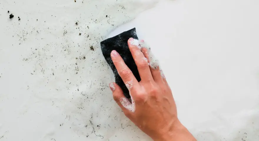 Main d'une personne lavant un mur humide avec une éponge savonneuse. Des saletés et des débris sont visibles sur la surface.