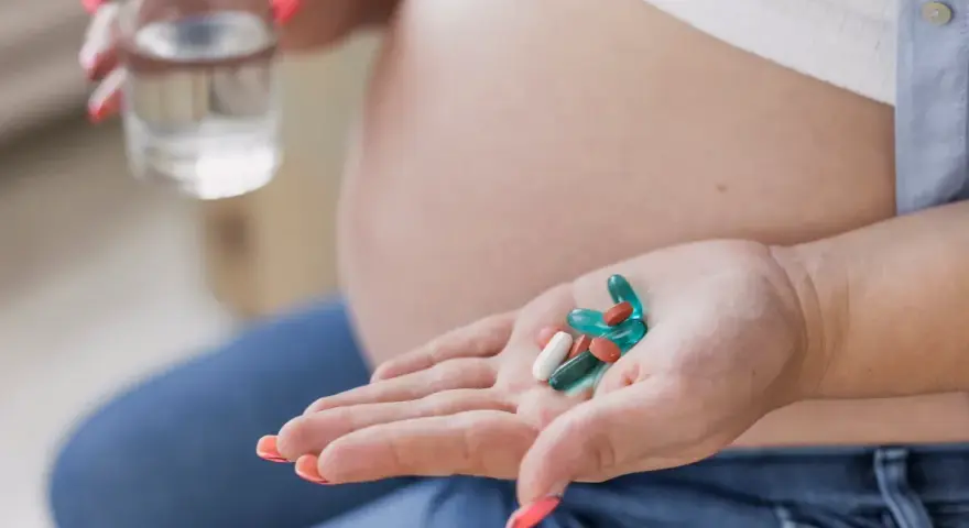 Kobieta w ciąży trzyma na dłoni suplementy, w tym żelazo, przygotowując się do ich zażycia z wodą.