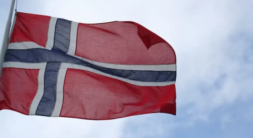 Flaga Norwegii powiewa na wietrze na tle błękitnego nieba z chmurami.