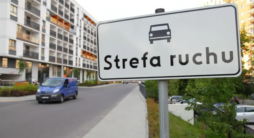 Znak "Strefa ruchu" przed nowoczesnym budynkiem mieszkalnym. Niebieski bus jedzie po drodze.