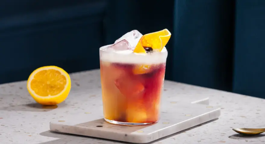 Orzeźwiający drink z whisky i cytryną, ozdobiony skórką cytrusową i lodem. Obok leży połówka cytryny.