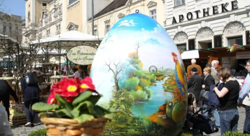 Großes, bemaltes Ei mit Frühlingslandschaft und Hahn, ein Osterbrunnen, der erklärt, warum ist Ostern immer wann anders.