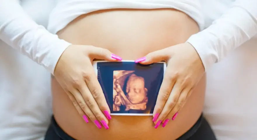 Ręce z różowymi paznokciami obejmują brzuch, na którym widnieje zdjęcie z 8 tygodnia ciąży usg 3d.