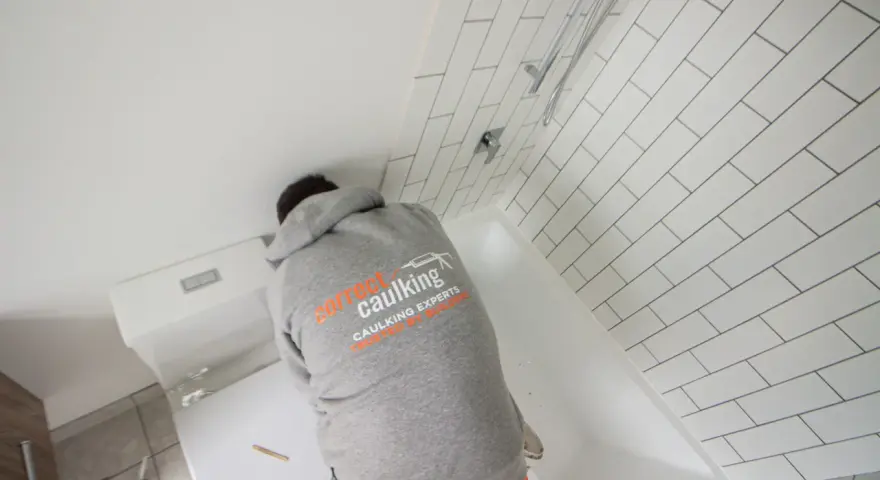 A person in a "Correct Caulking" hoodie works on a bathtub, demonstrating how to caulk a bathtub with white subway tiles in the background.