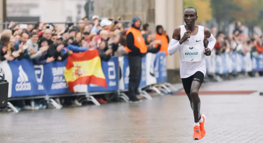 Eliud Kipchoge corre in una maratona, mostrando quanto è lunga una maratona. Pubblico festante e bandiera spagnola sullo sfondo.