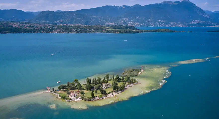 L'isola dei conigli sul Lago di Garda, un gioiello verde con case e alberi, circondata da acque turchesi e montagne sullo sfondo.