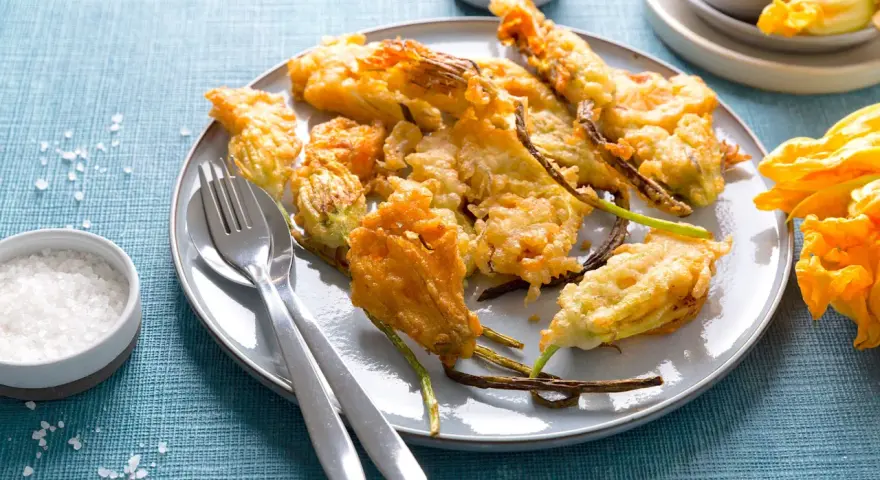 Piatto di fiori di zucca in pastella dorati e croccanti, serviti con sale grosso e posate.