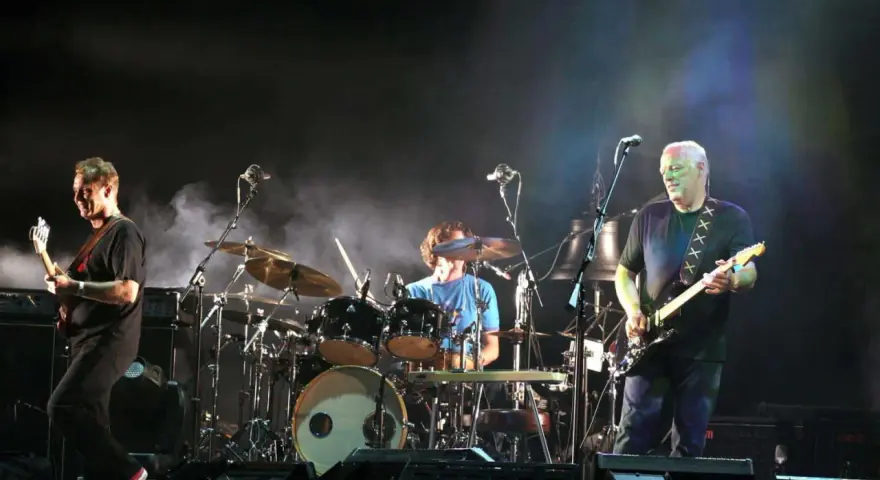 Les membres de Pink Floyd sur scène, David Gilmour à la guitare, Nick Mason à la batterie, et un autre musicien.
