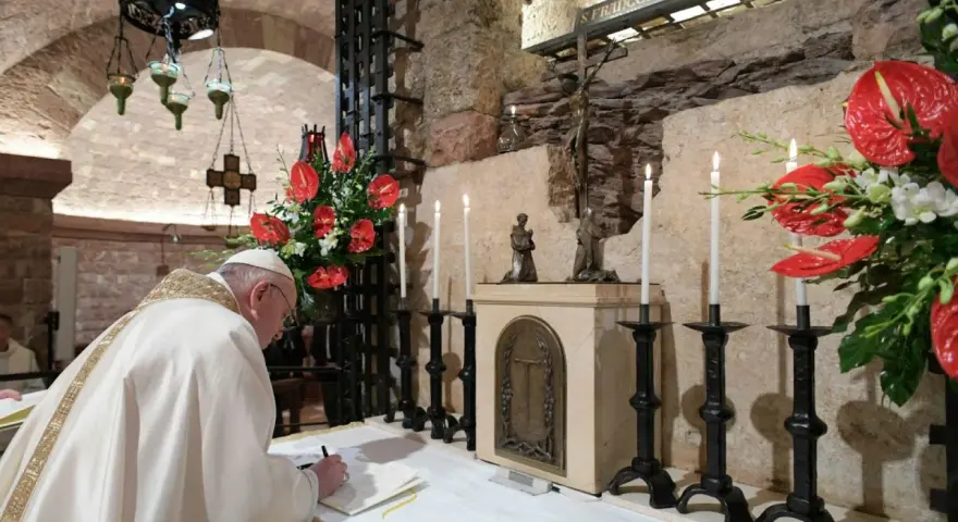 Papst Franziskus schreibt in einem Buch, während Kerzen und rote Anthurien brennen. Dies erinnert an die Bedeutung einer Enzyklika, eines wichtigen päpstlichen Schreibens.