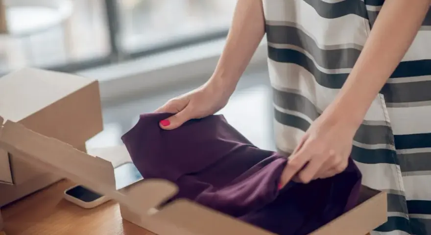 Kobieta pakuje ciemnofioletową sukienkę do kartonu, przygotowując ją do **vinted zwroty**.