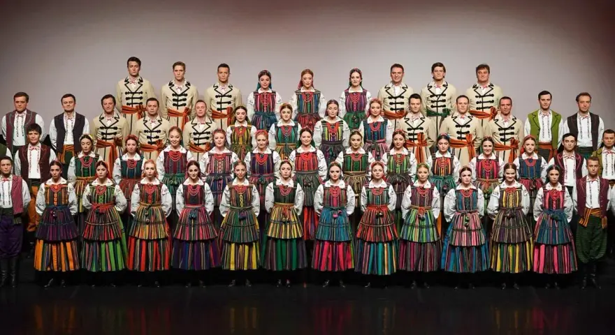 Zespół w barwnych strojach ludowych, prezentujący mazowieckie piosenki ludowe.