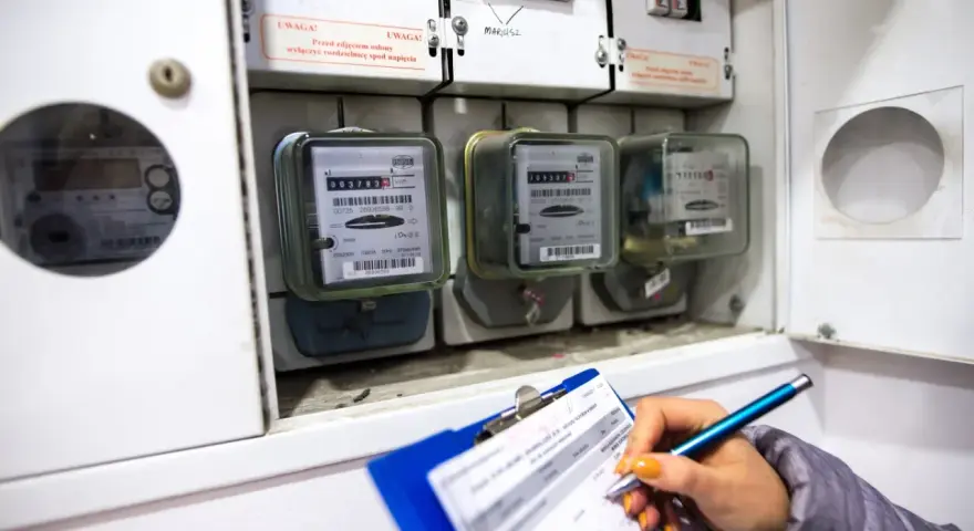Kontrola liczników energii elektrycznej. Ręka z długopisem zapisuje dane na kartce, obok widoczne liczniki i bezpieczniki.
