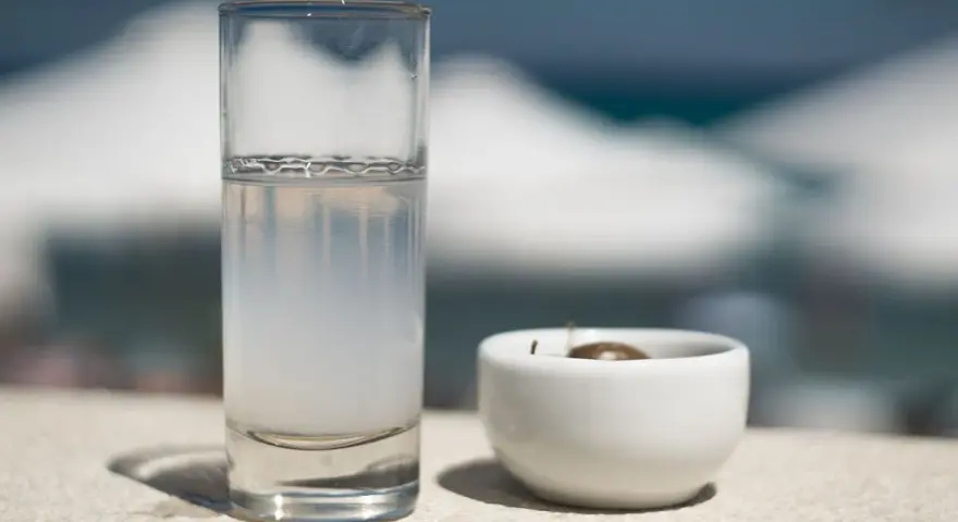 Vaso de pastis bebida, con una aceituna en un cuenco blanco. El fondo está desenfocado, sugiriendo un día soleado.