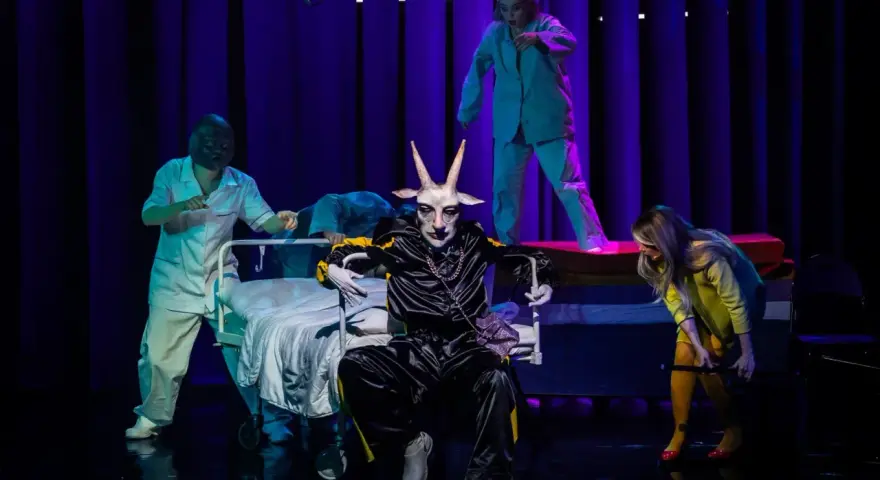 Na scenie, w surrealistycznej atmosferze, panna obsada w stroju z głową kozła siedzi na wózku. Wokół niej, w jasnych fartuchach, poruszają się postacie przypominające personel medyczny.
